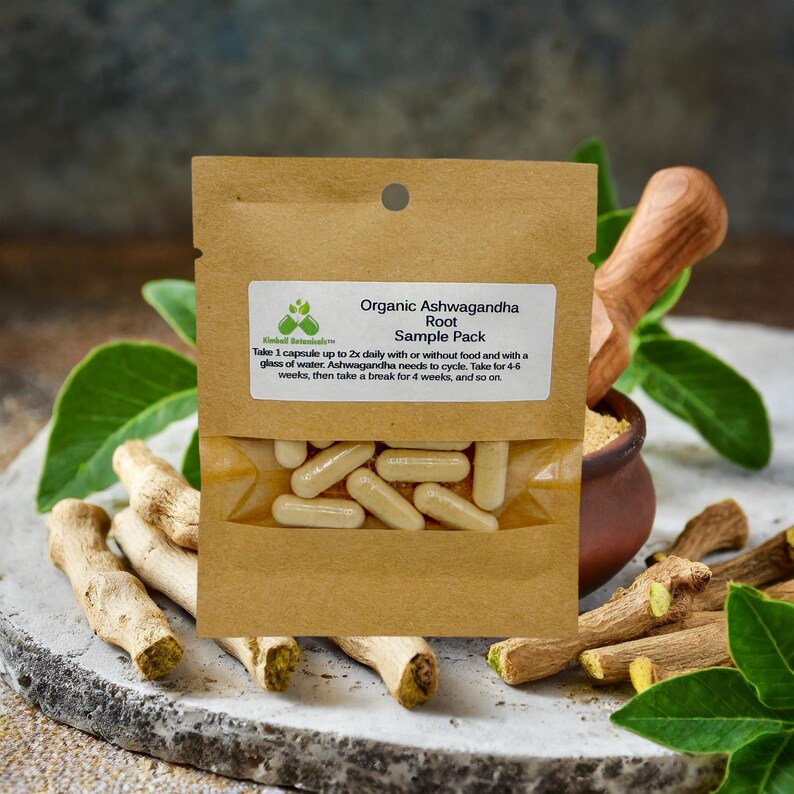 May include: A brown paper pouch with a white label that reads "Organic Ashwagandha Root Sample Pack". The pouch contains a sample pack of capsules. The pouch is sitting on a table with a mortar and pestle and some dried roots.
