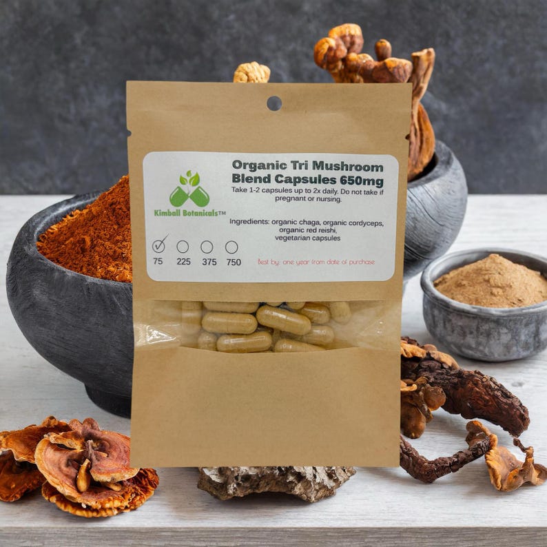 May include: A brown paper pouch with a clear window showing a pile of beige capsules. The pouch is labeled "Organic Tri Mushroom Blend Capsules 650mg" and lists the ingredients as organic chaga, organic cordyceps, organic red reishi, and vegetarian capsules. The pouch is sitting on a wooden surface with dried mushrooms.