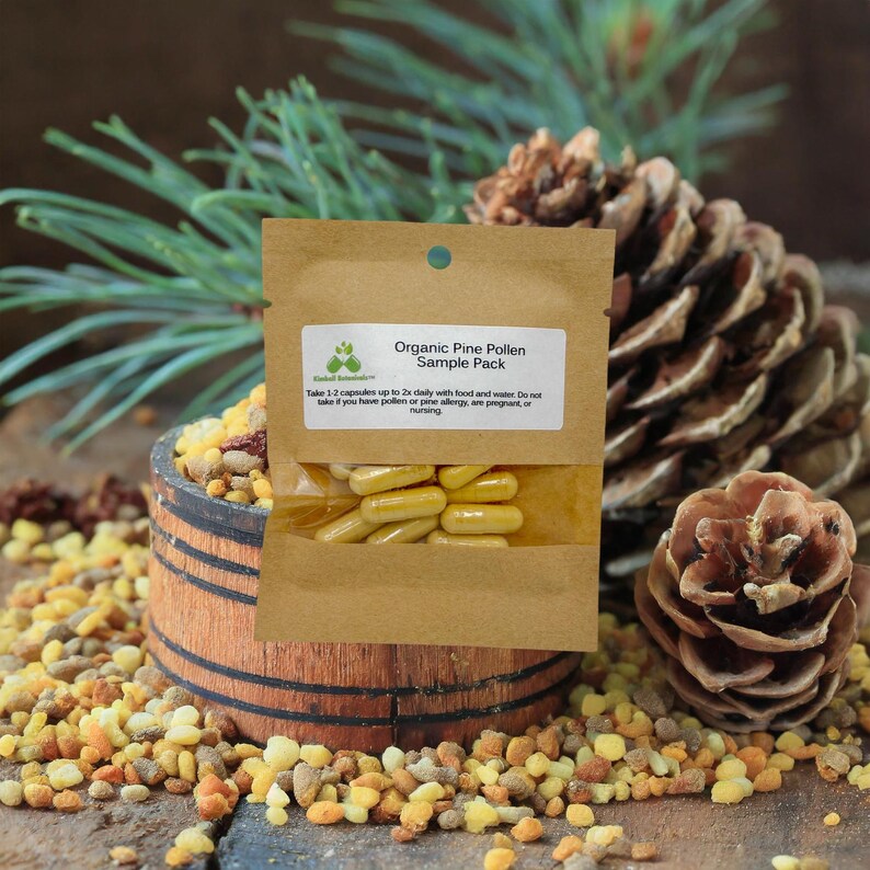 May include: A brown paper packet labeled "Organic Pine Pollen Sample Pack" containing yellow capsules. The packet rests on a wooden barrel surrounded by pine cones and small, colorful granules. Green pine needles are in the background.