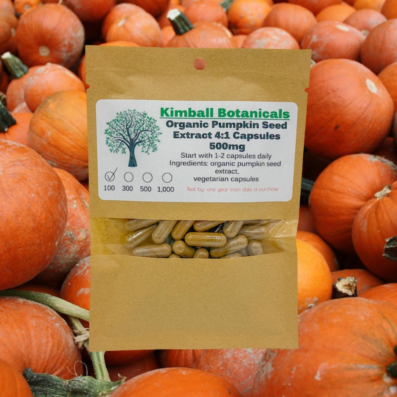 May include: A brown paper bag with a label that reads "Kimball Botanicals Organic Pumpkin Seed Extract 4:1 Capsules 500mg". The bag contains a clear plastic bag filled with brown capsules. The bag is surrounded by a pile of orange pumpkins.