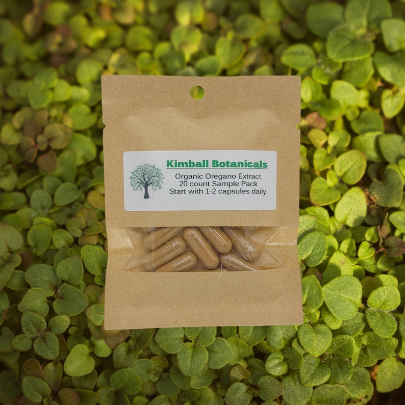 May include: A brown paper packet of Kimball Botanicals Organic Oregano Extract capsules. The packet has a clear window showing the capsules and a label with the brand name and product information. The background is a bed of green leaves.
