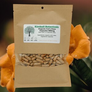 Pu&ograve; includere: Un sacchetto di carta marrone con un'etichetta che recita "Kimball Botanicals Organic Cat's Claw Capsules 500mg Start with 1-2 capsules daily Ingredients: organic cat's claw, vegetarian capsules". Il sacchetto &egrave; pieno di capsule e si trova su un fiore giallo.