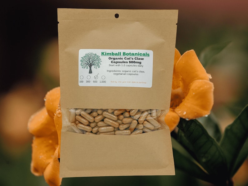 Pu&ograve; includere: Un sacchetto di carta marrone con un'etichetta che recita "Kimball Botanicals Organic Cat's Claw Capsules 500mg Start with 1-2 capsules daily Ingredients: organic cat's claw, vegetarian capsules". Il sacchetto &egrave; pieno di capsule e si trova su un fiore giallo.