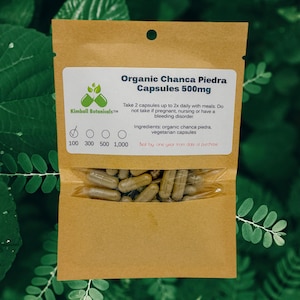 May include: A brown paper pouch with a label that reads "Organic Chanca Piedra Capsules 500mg". The label also includes instructions for use and ingredients. The pouch contains a clear plastic bag filled with brown capsules.