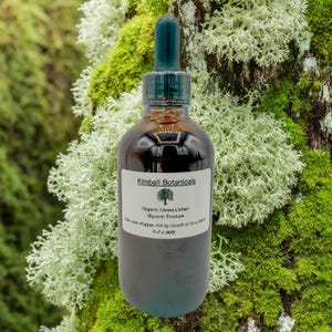 May include: A dark amber glass bottle with a dropper, labeled "Kimball Botanicals." The label reads "Organic Usnea Lichen Glycerin Tincture." The bottle is set against a backdrop of green moss and white lichen. The text on the label also says "Take one dropper full by mouth or in a drink 1-2 x daily."