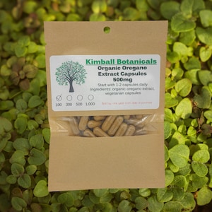 May include: A brown paper packet of Kimball Botanicals Organic Oregano Extract Capsules 500mg. The label includes a tree graphic and text detailing usage and ingredients. The packet contains brown capsules and is set against a green leafy background.