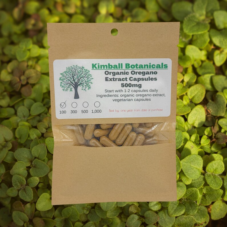 May include: A brown paper packet of Kimball Botanicals Organic Oregano Extract Capsules 500mg. The label includes a tree graphic and text detailing usage and ingredients. The packet contains brown capsules and is set against a green leafy background.