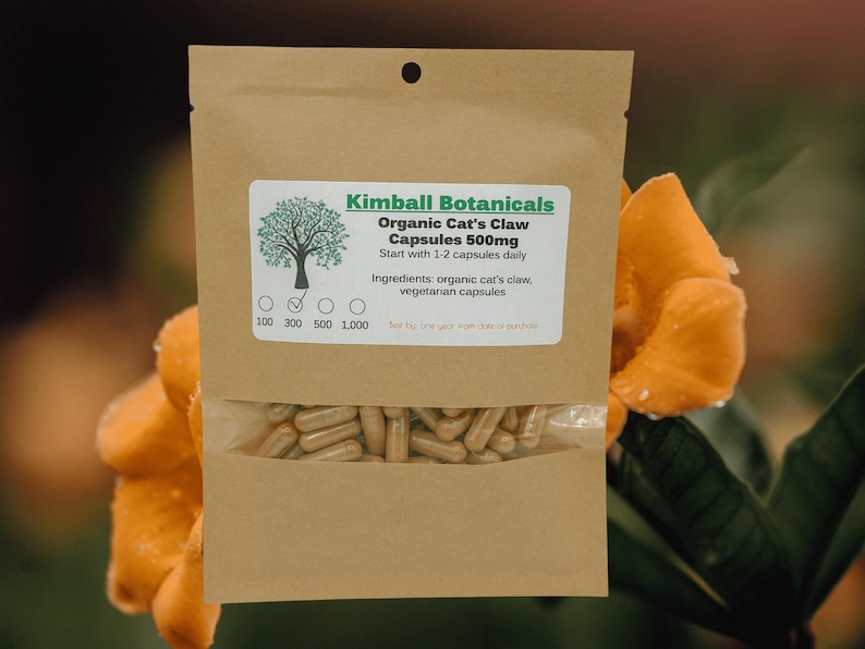 Pu&ograve; includere: Un sacchetto di carta marrone con un'etichetta bianca che recita "Kimball Botanicals Organic Cat's Claw Capsules 500mg Start with 1-2 capsules daily Ingredients: organic cat's claw, vegetarian capsules 100 300 500 1,000". Il sacchetto contiene capsule.