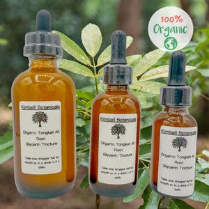 May include: Three amber glass bottles with droppers, labeled "Kimball Botanicals Organic Tongkat Ali Root Glycerin Tincture". The bottles are arranged in a row from largest to smallest, with the largest bottle in the front. The bottles are sitting on a green leafy background. The text "100% Organic" is in a white circle with a green globe icon.