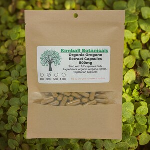 May include: A brown paper bag of Kimball Botanicals Organic Oregano Extract Capsules, 500mg. The bag has a clear window showing the capsules. The label includes the text "Start with 1-2 capsules daily" and "vegetarian capsules". The background is green foliage.