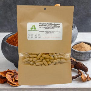 May include: A brown paper bag with a clear plastic window showing a large quantity of brown capsules. The bag is labeled "Organic Tri Mushroom Blend Capsules 650mg" and lists the ingredients as organic chaga, organic cordyceps, organic red reishi, and vegetarian capsules. The bag also has a label with the Kimball Botanical logo and a size chart with the numbers 75, 225, 375, and 750.