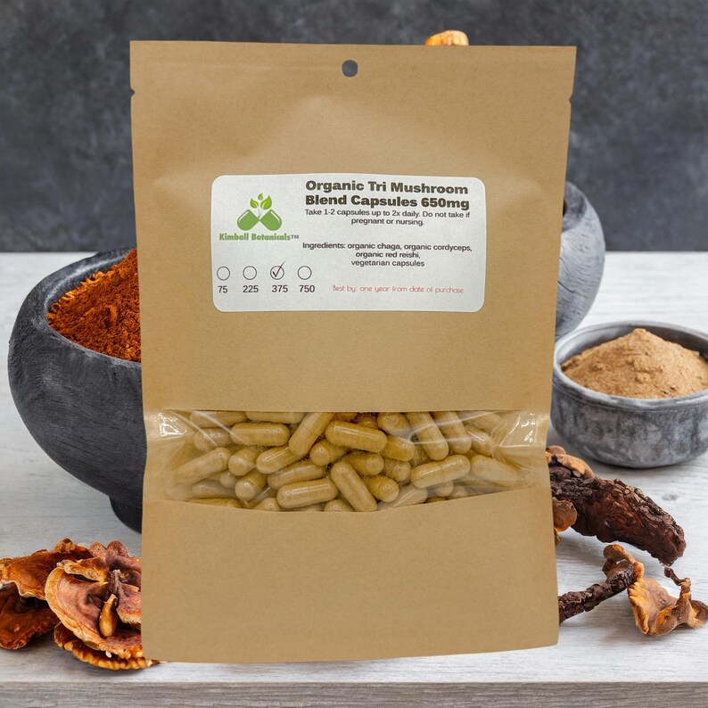 May include: A brown paper bag with a clear plastic window showing a large quantity of brown capsules. The bag is labeled "Organic Tri Mushroom Blend Capsules 650mg" and lists the ingredients as organic chaga, organic cordyceps, organic red reishi, and vegetarian capsules. The bag also has a label with the Kimball Botanical logo and a size chart with the numbers 75, 225, 375, and 750.
