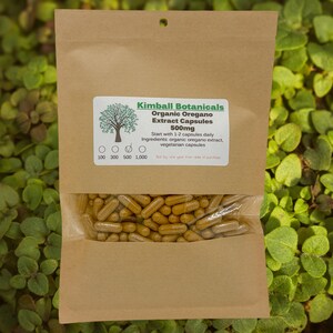 May include: A brown paper bag with a clear window, filled with yellow-brown capsules. The bag has a white label with the text "Kimball Botanicals Organic Oregano Extract Capsules 500mg". The label also includes the text "Start with 1-2 capsules daily".