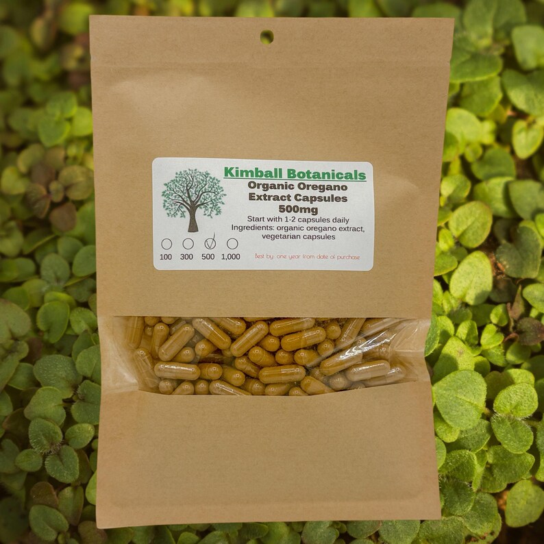 May include: A brown paper bag with a clear window, filled with yellow-brown capsules. The bag has a white label with the text "Kimball Botanicals Organic Oregano Extract Capsules 500mg". The label also includes the text "Start with 1-2 capsules daily".