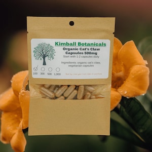 Pu&ograve; includere: Una busta di carta marrone con un'etichetta che recita "Kimball Botanicals Organic Cat's Claw Capsules 500mg". La busta contiene un mucchio di capsule marroni. La busta &egrave; appoggiata su un fiore giallo.