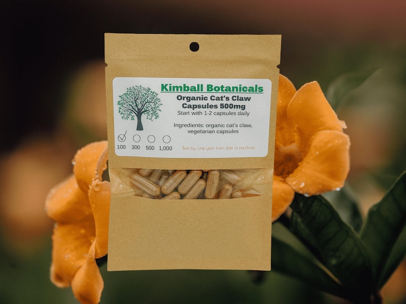 Pu&ograve; includere: Una busta di carta marrone con un'etichetta che recita "Kimball Botanicals Organic Cat's Claw Capsules 500mg". La busta contiene un mucchio di capsule marroni. La busta &egrave; appoggiata su un fiore giallo.