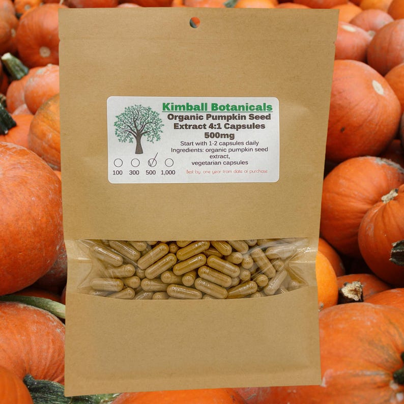 May include: A brown paper bag with a label that reads "Kimball Botanicals Organic Pumpkin Seed Extract 4:1 Capsules 500mg". The bag contains a clear plastic bag filled with brown capsules. The bag is sitting on a pile of orange pumpkins.