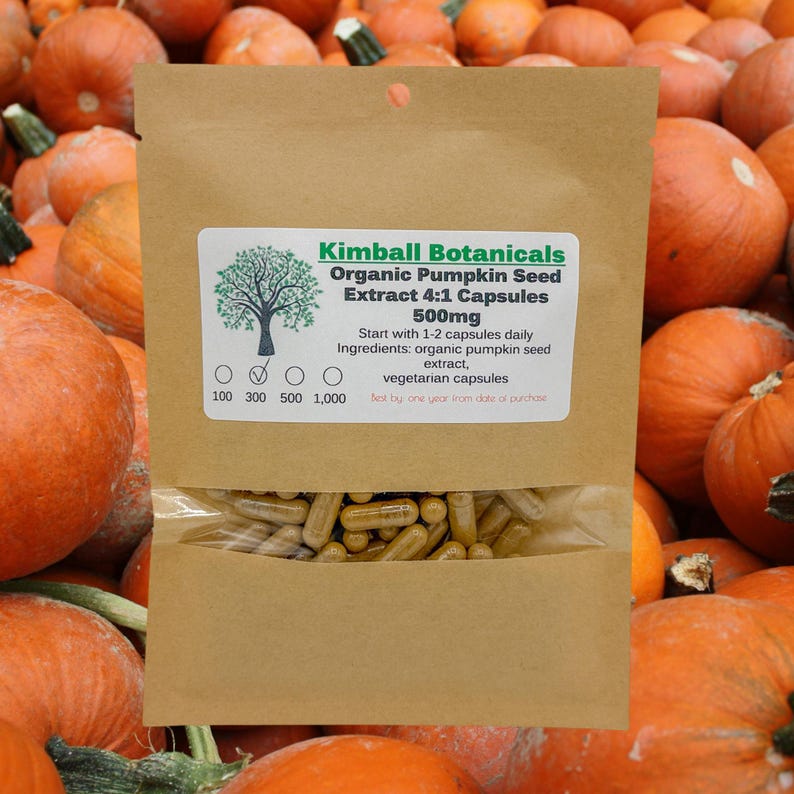May include: A brown paper bag with a label that reads "Kimball Botanicals Organic Pumpkin Seed Extract 4:1 Capsules 500mg". The bag contains a clear plastic bag filled with brown capsules. The bag is sitting on a pile of orange pumpkins.