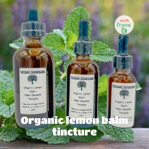 May include: Three brown glass bottles with droppers, labeled "Organic Lemon Balm Tincture." The bottles have white labels with a tree logo and the text "Kimball Botanicals." The image includes the text "Organic lemon balm tincture" at the bottom and a "100% Organic" sticker.