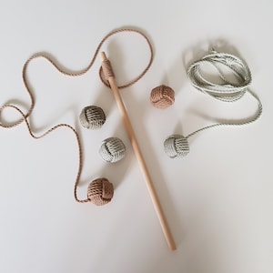 Pode incluir: Um conjunto de bolas de corda atadas em bege e verde, com um pau de madeira e um pedaço de corda bege. As bolas estão amarradas à corda e podem ser usadas para uma variedade de jogos.