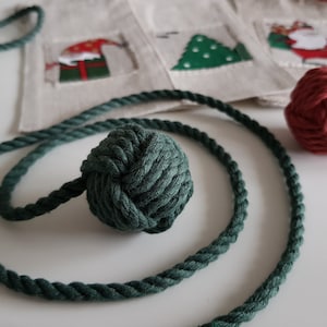 Könnte beinhalten: Ein grünes Seil mit einer geknoteten Kugel am Ende. Das Seil ist auf einer weißen Oberfläche aufgewickelt. Im Hintergrund befindet sich eine rote, geknotete Kugel.