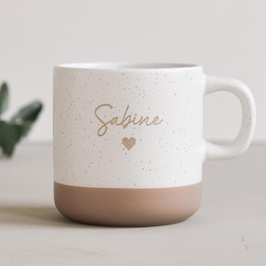 Op de afbeelding: Witte keramische mok met een gespikkeld ontwerp en een bruine basis. Het woord "Sabine" is in gouden letters geschreven, met daaronder een klein gouden hartje. De mok heeft een gebogen handvat.