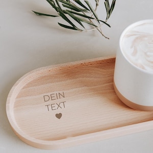 Könnte beinhalten: Eine weiße Keramiktasse mit beigem Boden, gefüllt mit einem schaumigen Getränk, steht auf einem ovalen Tablett aus hellem Holz. Das Tablett trägt den Text "DEIN TEXT" und ein Herzsymbol. Ein Zweig Grünzeug befindet sich im Hintergrund.
