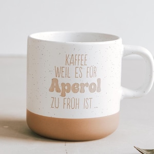 Könnte beinhalten: Weißer Keramikbecher mit gesprenkelter Oberfläche und braunem Boden. Der Becher trägt den Text "KAFFEE WEIL ES FÜR APEROL ZU FRÜH IST..." in braunen Buchstaben. Ein tolles Geschenk für Kaffee- und Aperol-Liebhaber.
