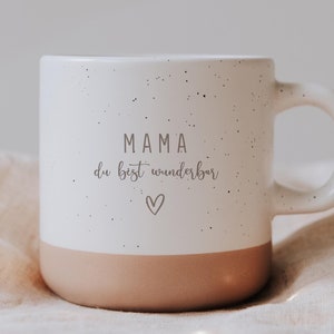Op de afbeelding: Witte keramische mok met een gespikkelde afwerking en een bruine basis. De mok heeft de tekst "MAMA du bist wunderbar" en een hartsymbool. Ontworpen voor warme of koude dranken.