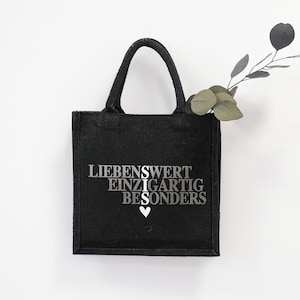 Könnte beinhalten: Schwarze Tragetasche mit weißem Text, der "LIEBENSWERT EINZIGARTIG BESONDERS" mit einem kleinen weißen Herzsymbol lautet.