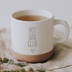 Könnte beinhalten: Weißer Keramikbecher mit gesprenkeltem Finish und braunem Boden. Der Becher ist mit einer hellbraunen Flüssigkeit gefüllt und trägt den Text "HOT DAD CLUB" mit einem Herz. Ein Zweig getrockneten Grüns liegt auf beigem Stoff.