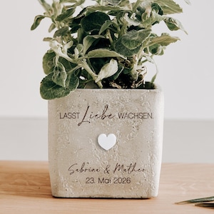 May include: A square, light gray concrete planter with a small green plant. The planter has the words "LASST LIEBE WACHSEN" and the names "Sabrina & Mathias" with the date "23. Mai 2026" printed on it. A white heart is also on the planter.