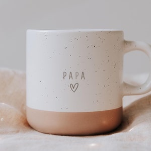 Peut inclure: Une tasse en céramique blanche avec un motif tacheté et une base pêche. Le mot "PAPA" et un cœur sont imprimés sur le devant. La tasse est posée sur un tissu beige.