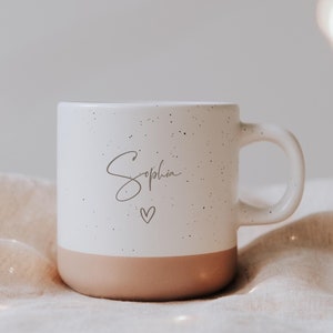 Könnte beinhalten: Weiß gesprenkelter Keramikbecher mit pfirsichfarbenem Boden. Der Becher trägt den Namen "Sophia" in Schreibschrift sowie ein kleines Herz. Der Becher hat einen Henkel und wird wahrscheinlich für Getränke verwendet.