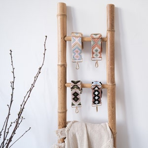 Puede incluir: Cuatro correas de colores con patrones y herrajes dorados colgando de una escalera de bambú. Las correas están hechas de tejido tejido y tienen un diseño geométrico. Una manta beige está drapeada sobre el peldaño inferior de la escalera.