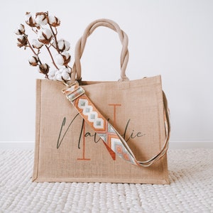 May include: A natural jute tote bag with a personalised name "Natalie" printed in black script. The bag has a long, woven strap with a geometric pattern in orange, grey, and white.