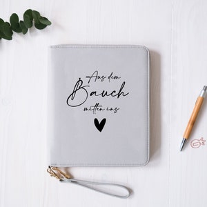 Può includere: Un diario rilegato in pelle grigia con una scritta nera che recita "Aus dem Bauch mitten ins" con un piccolo cuore nero. Il diario ha una cerniera dorata e un cinturino grigio.