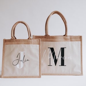 Könnte beinhalten: Zwei personalisierte Jute-Tragetaschen. Die größere Tasche hat ein schwarzes "M" für "Marion" und die kleinere Tasche ein weißes "J" für "Julia".