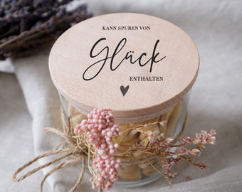 Könnte beinhalten: Ein Glasgefäß mit einem Holzdeckel, auf dem "Kann Spuren von Glück enthalten" steht. Das Gefäß ist mit getrockneten Blumen gefüllt und mit einer Schnur gebunden.