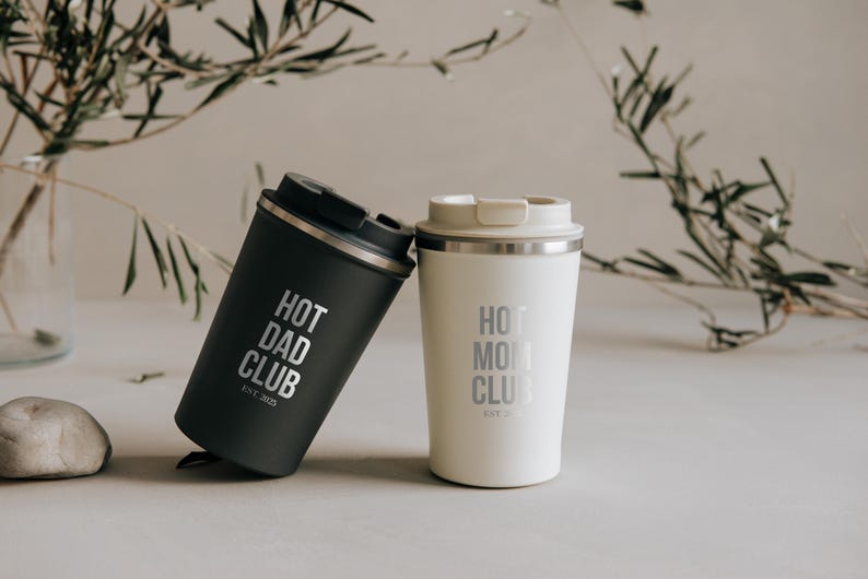 Zwei stilvolle Thermobecher, einer in elegantem Schwarz mit der Aufschrift "Hot Dad Club" und der andere in edlem Weiß mit "Hot Mom Club". Perfekt für Eltern, die ihren Kaffee heiß und ihren Stil cool halten möchten. Ein ideales Geschenk, das sowohl praktisch als auch emotional berührt. Im Hintergrund dezente Pflanzen, die eine harmonische Atmosphäre schaffen.