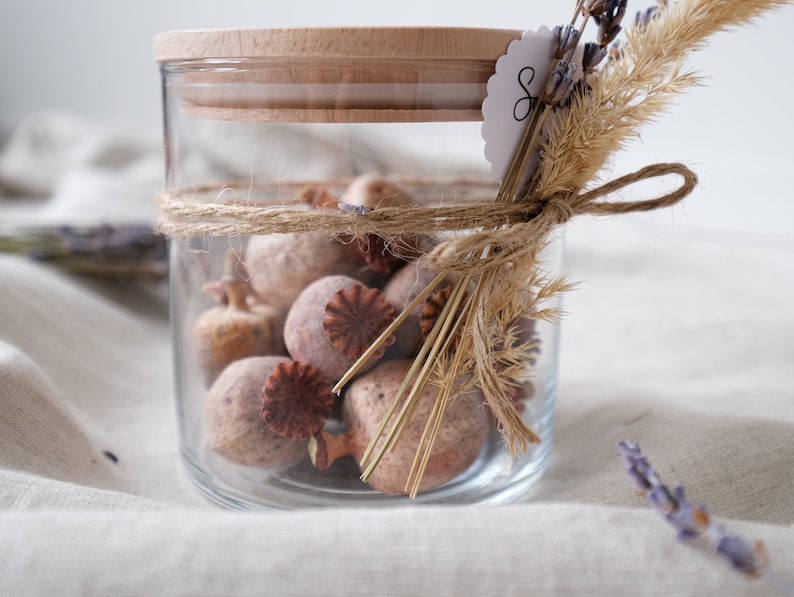 Hochwertiges Glasgefäß mit Holzdeckel, gefüllt mit natürlichem Potpourri. Dekoratives Seil umschließt das Glas, kombiniert mit zarten, getrockneten Blumen. Perfekt für eine gemütliche Atmosphäre in Ihrem Zuhause. Ideal als stilvolles Geschenk oder als elegantes Dekorelement, das Wärme und Naturverbundenheit ausstrahlt.