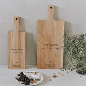 K&ouml;nnte beinhalten: Zwei Holzschneidebretter mit Griffen, eines gro&szlig; und eines klein. Die Bretter tragen den Text "HAND IN HAND" und andere Texte. Ein Teller mit Essen und eine kleine Pflanze befinden sich im Vordergrund.