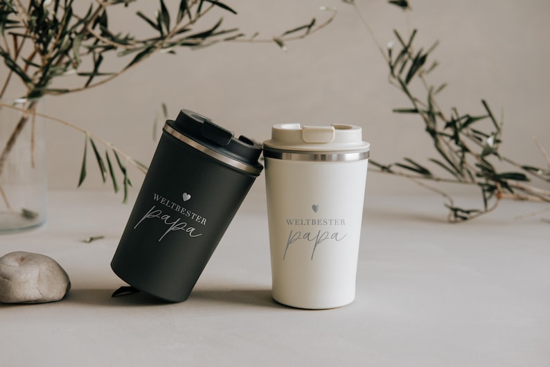 Zwei stylische Thermobecher mit der Aufschrift "Weltbester Papa" stehen auf einem Tisch. Der eine Becher ist in elegantem Schwarz, der andere in modernem Weiß gestaltet. Diese Becher sind das perfekte Geschenk, um Wertschätzung und Wärme auszudrücken. Ideal für jeden Vater, der seinen Kaffee unterwegs genießen möchte und stets an die Liebe seiner Familie erinnert werden will.
