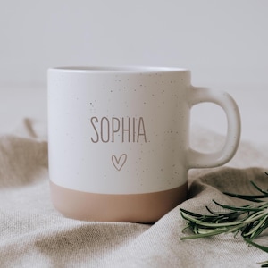 Può includere: Tazza in ceramica bianca con il nome "SOPHIA" stampato in grigio e un piccolo simbolo a forma di cuore. Il fondo della tazza è rosa chiaro. La tazza ha un manico ed è macchiata di piccoli puntini neri.