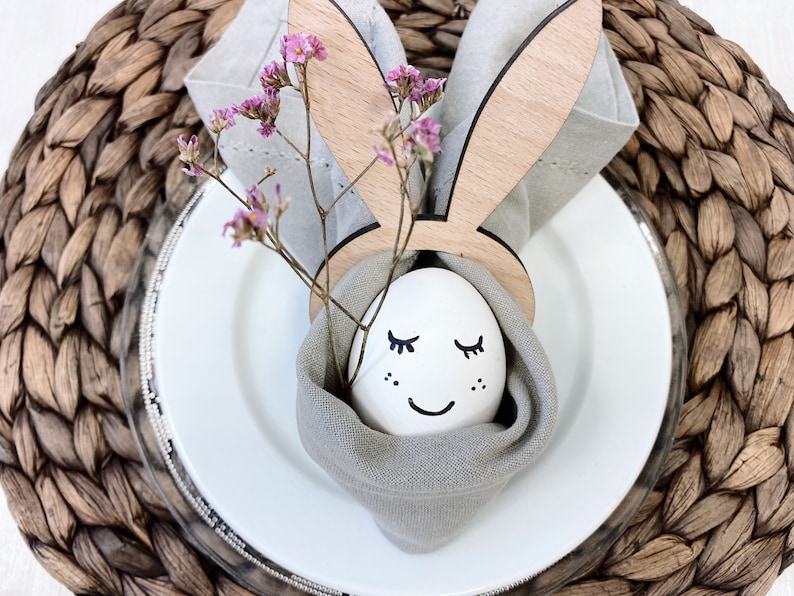 Osterdeko-Set: Handgefertigter Eierbecher aus Holz in Hasenform, ergänzt durch ein liebevoll bemaltes Ei mit Gesicht. Eingehüllt in feines Leinen, wird es von zarten, violetten Blumen geschmückt. Perfekt für eine frühlingshafte Tischdekoration, die Wärme und Freude in Ihr Zuhause bringt. Ideal für Ostern oder als Geschenkidee.