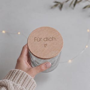 Peut inclure: Un bocal en verre transparent avec un couvercle circulaire en bois brun clair. Le couvercle porte l'inscription gravée "Für dich." accompagnée d'un petit symbole de cœur. Le bocal est cylindrique avec des parois en verre lisse.