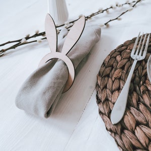 Elegantes Tischset mit natürlichem Flair: Ein stilvoller Serviettenring in Hasenform aus Holz umschließt eine beige Stoffserviette. Daneben ein handgeflochtener Platzdeckchen und edles, graviertes Glasgeschirr. Ideal für eine festliche Ostertafel, die Wärme und Gemütlichkeit ausstrahlt. Perfekt, um Ihre Gäste zu begeistern und den Tisch in ein harmonisches Gesamtbild zu verwandeln.