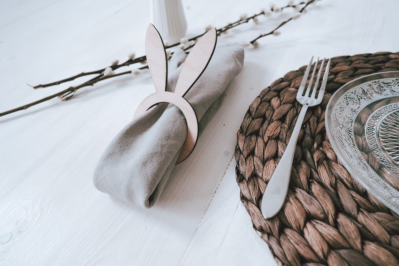 Elegantes Tischset mit natürlichem Flair: Ein stilvoller Serviettenring in Hasenform aus Holz umschließt eine beige Stoffserviette. Daneben ein handgeflochtener Platzdeckchen und edles, graviertes Glasgeschirr. Ideal für eine festliche Ostertafel, die Wärme und Gemütlichkeit ausstrahlt. Perfekt, um Ihre Gäste zu begeistern und den Tisch in ein harmonisches Gesamtbild zu verwandeln.