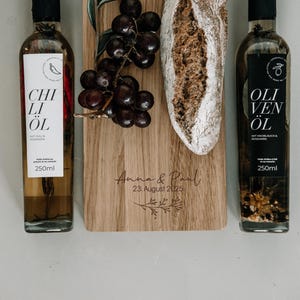 Può includere: Una natura morta con due bottiglie di olio da 250 ml, una con l'etichetta "Chili Öl" e l'altra "Oliven Öl", che affiancano un tagliere in legno con uva e una baguette. La tavola è incisa con un nome e una data.