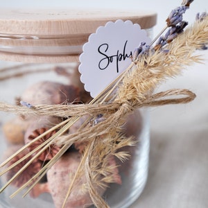 Liebevoll gestaltetes Glas mit Holzdeckel, gefüllt mit duftendem Potpourri und dekoriert mit natürlichen Gräsern und Lavendel. Perfekt als personalisiertes Geschenk oder stilvolle Deko. Der handgeschriebene Namensanhänger und die zarte Juteschnur verleihen dem Arrangement eine persönliche Note und versprühen einen Hauch von ländlicher Eleganz. Ideal für besondere Anlässe.
